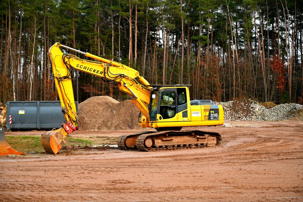 A yellow excavator at work in a forested construction site, surrounded by dirt and gravel.